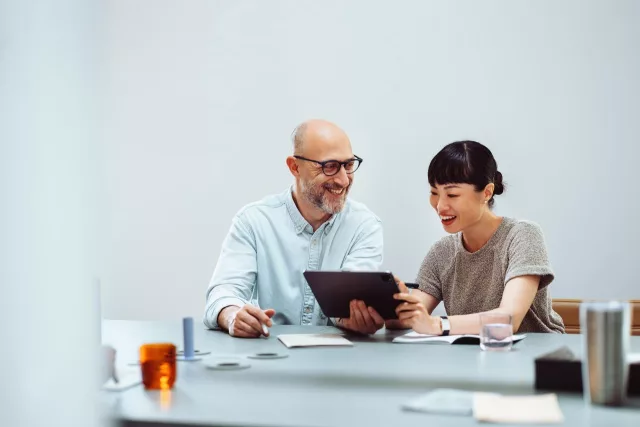Das Bild zeigt eine Frau und einen Mann, die an einem Tisch sitzen und gemeinsam an einem Tablet arbeiten. Auf dem Tisch liegen verschiedene Gegenstände wie Notizbücher, Stifte und ein Glas Wasser. Die Umgebung wirkt modern und aufgeräumt.
