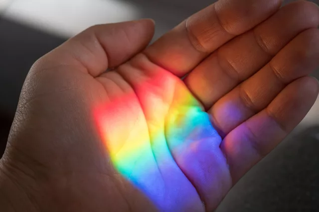 Eine Nahaufnahme einer Hand, die von einem bunten Lichtstrahl erleuchtet wird. Das Licht bildet ein Regenbogenmuster auf der Haut, das die Farben Rot, Orange, Gelb, Grün, Blau und Violett umfasst. Die Hand ist leicht geöffnet und die Finger sind entspannt. Der Hintergrund ist unscharf, wodurch der Fokus auf die Hand und das faszinierende Farbenspiel gelegt wird. Das Bild vermittelt eine ruhige und magische Atmosphäre, die die Schönheit und Vielfalt des Lichts einfängt.