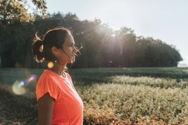 Eine Frau steht in einem offenen Feld und genießt die Natur. Sie trägt ein leuchtend orangefarbenes T-Shirt und hat ihre Haare zu einem lockeren Pferdeschwanz gebunden. Die Sonne scheint durch die Bäume im Hintergrund und erzeugt einen warmen Lichtschein, der die Szene idyllisch und friedlich wirken lässt. Die Umgebung ist grün und weitläufig, mit einem Wald im Hintergrund, der eine ruhige und entspannende Atmosphäre schafft.