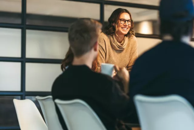 Das Bild zeigt drei Personen in einem modernen Büroraum, die miteinander sprechen. Zwei Männer sitzen an einem Konferenztisch, während die Frau am Tischende steht. Im Hintergrund sind große Fenster mit Milchglas zu sehen.