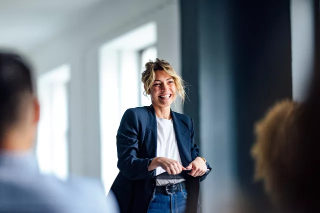 Eine Person hält einen Vortrag oder eine Präsentation vor mehreren Zuhörern in einem modern eingerichteten Raum. Sie trägt einen dunkelblauen Blazer, ein weißes Oberteil und eine Jeans. Die Atmosphäre wirkt professionell und aufmerksam.
