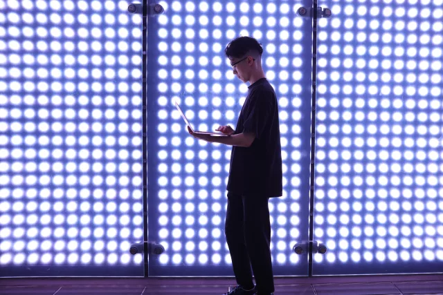 Eine Person steht seitlich vor einer leuchtenden Wand aus vielen LED-Lichtern und arbeitet an einem aufgeklappten Laptop. Das futuristisch wirkende Setting lässt den gesamten Hintergrund in hellem Weiß erstrahlen. Die dunkle Kleidung der Person hebt sich stark vom strahlenden Licht ab.