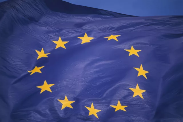 Eine blaue Fahne mit zwölf goldenen Sternen ist zu sehen und weht im Wind. Die Sterne sind im Kreis angeordnet und stehen für Einheit und Solidarität. Es handelt sich um die Flagge der Europäischen Union.