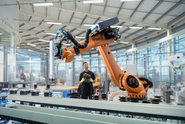 In einer modernen Fabrikhalle ist ein großer Industrieroboter im Einsatz. Im Hintergrund überwacht eine Person in Arbeitskleidung den Produktionsprozess über ein Steuergerät. Die Umgebung wirkt hell und technisch hochwertig, mit zahlreichen Glasabtrennungen und Maschinen.
