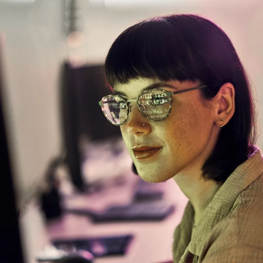 Auf dem Bild ist eine Frau zu sehen, die am Computer arbeitet. In ihrer Brille spiegeln sich Zahlenreihen vom Bildschirm des Computers.