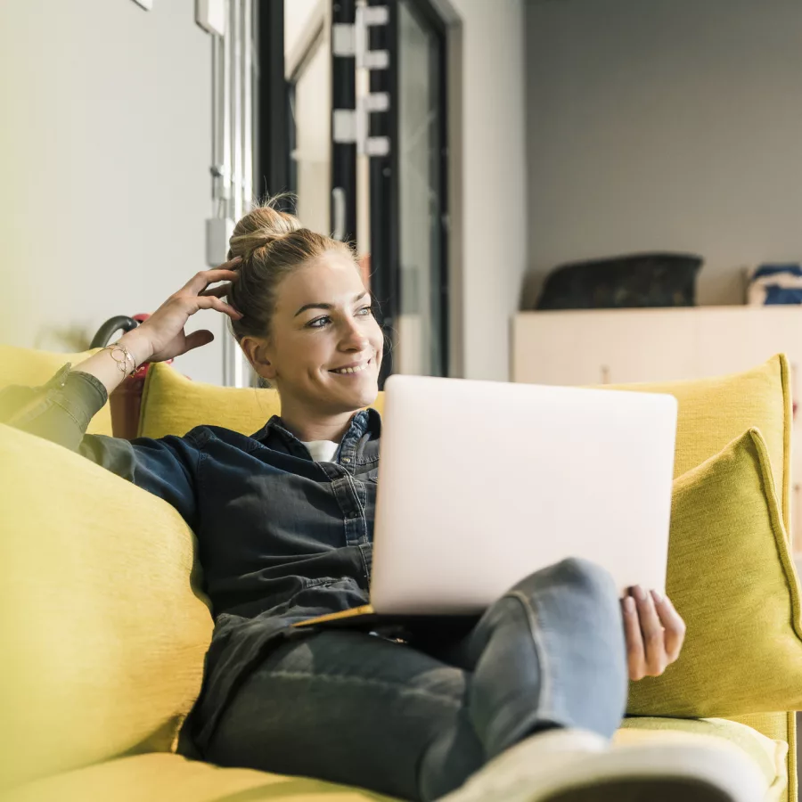 Auf dem Bild ist eine Frau zu sehen, die mit einem Laptop auf dem Schoß auf einem gelben Sofa sitz und lächelnd zur Seite schaut.