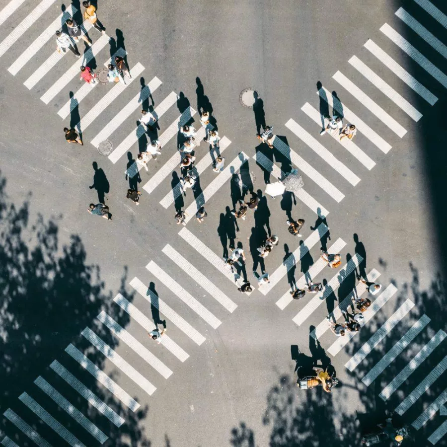 Das Bild zeigt eine Luftaufnahme einer Kreuzung mit Zebrastreifen in X-Form. Zahlreiche Menschen bewegen sich über die Straße, beleuchtet von einfallendem Licht.