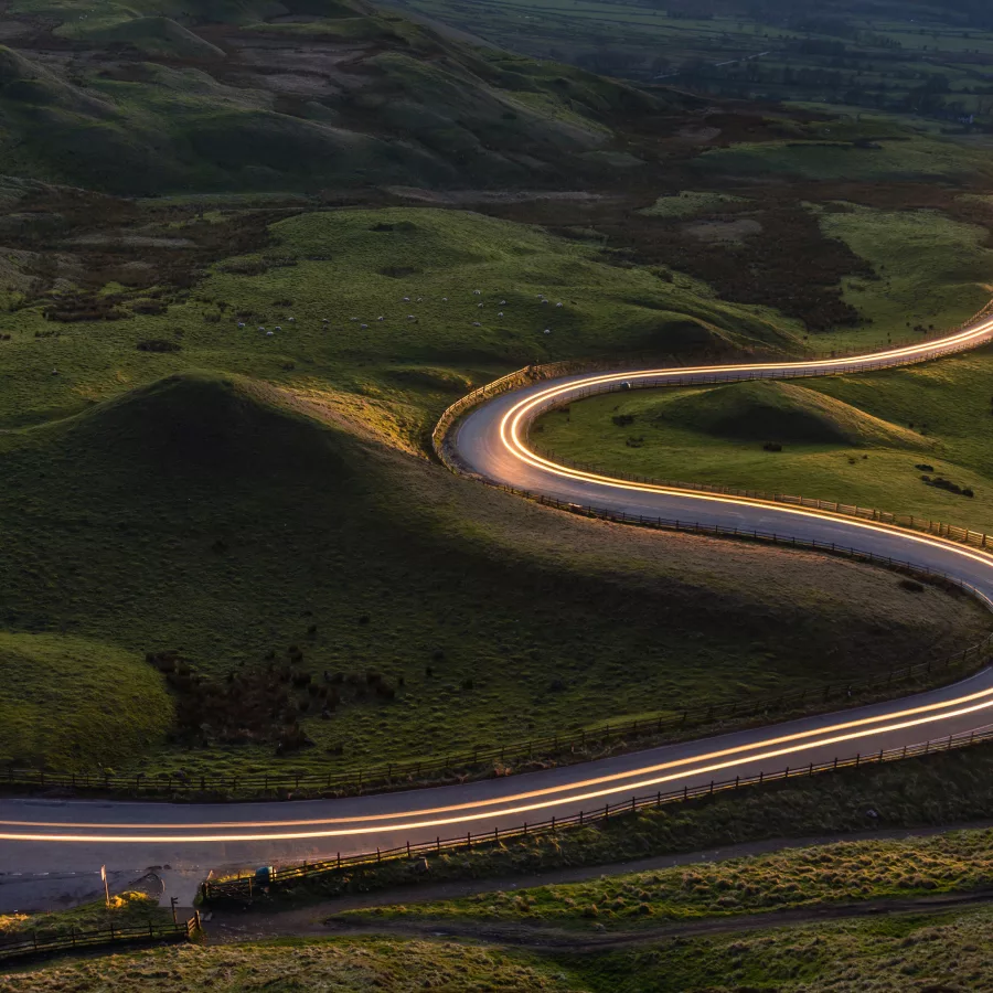 Eine kurvenreiche Straße schlängelt sich durch eine grüne, hügelige Landschaft bei Dämmerung, beleuchtet von den Lichtern vorbeifahrender Autos.