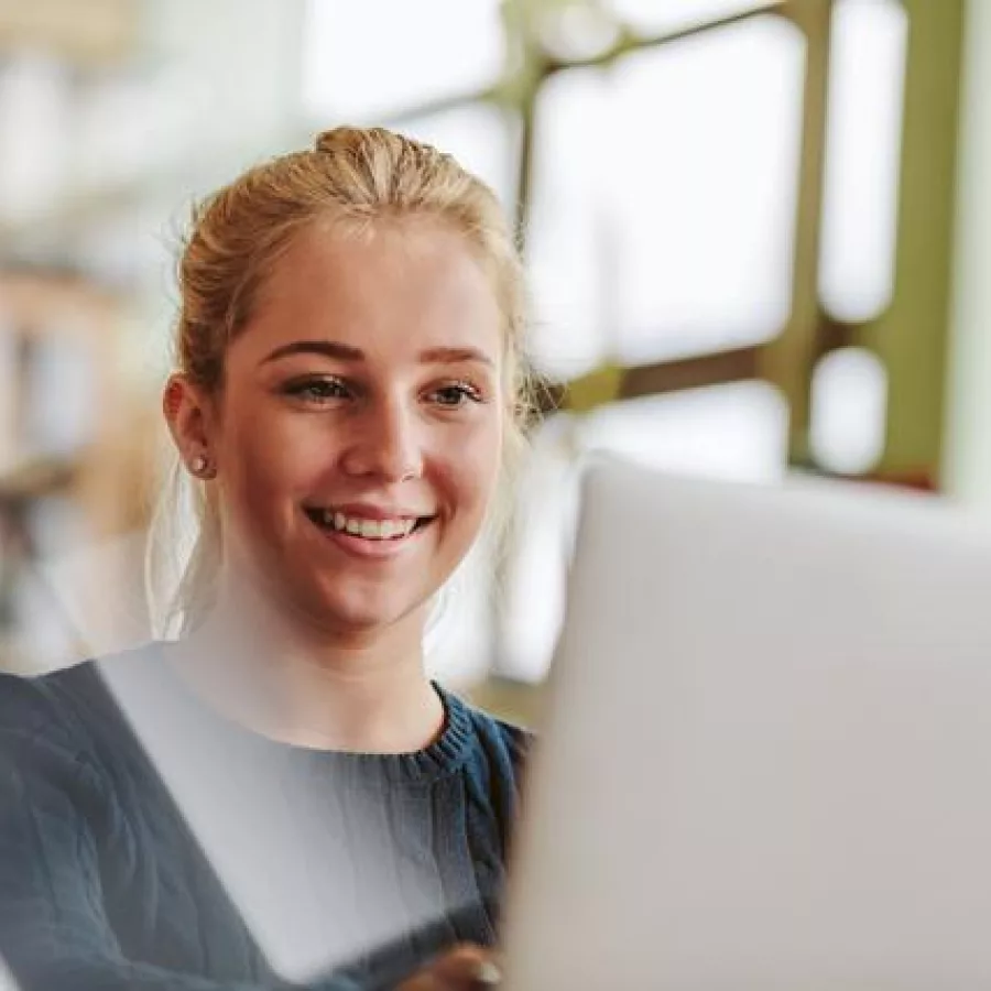 Das Bild zeigt eine junge Frau, die an einem Laptop arbeitet. Durch die großen Fenster im Hintergrund ist die Umgebung hell und gut beleuchtet.