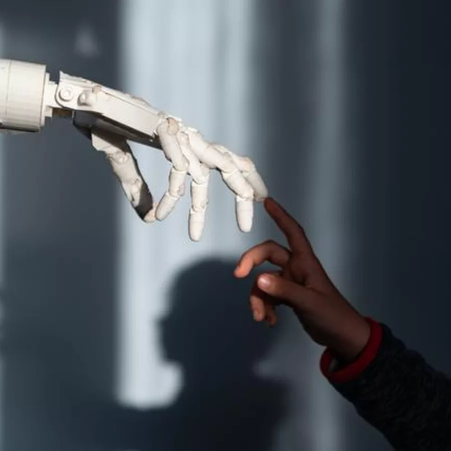 Das Bild zeigt eine menschliche Hand und eine Roboterhand, die sich fast berühren. Die menschliche Hand gehört einem schwarzen Kind, während die Roboterhand weiß und mechanisch ist, wodurch ein starker Farbkontrast entsteht. Durch das einfallende Licht entstehen Schatten im Hintergrund, die der Darstellung eine dramatische und symbolische Atmosphäre verleihen.