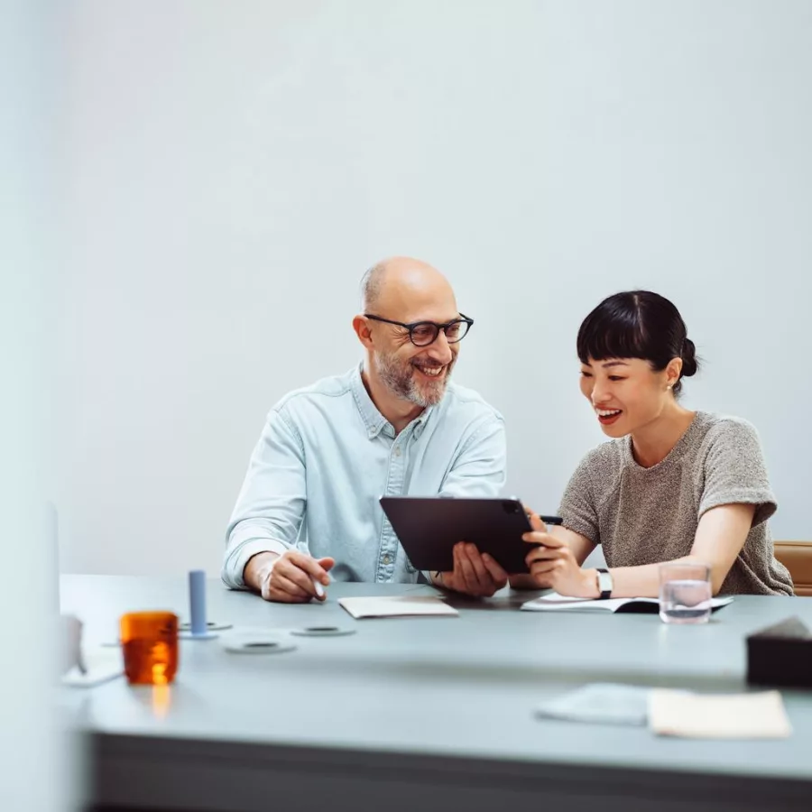 Das Bild zeigt eine Frau und einen Mann, die an einem Tisch sitzen und gemeinsam an einem Tablet arbeiten. Auf dem Tisch liegen verschiedene Gegenstände wie Notizbücher, Stifte und ein Glas Wasser. Die Umgebung wirkt modern und aufgeräumt.