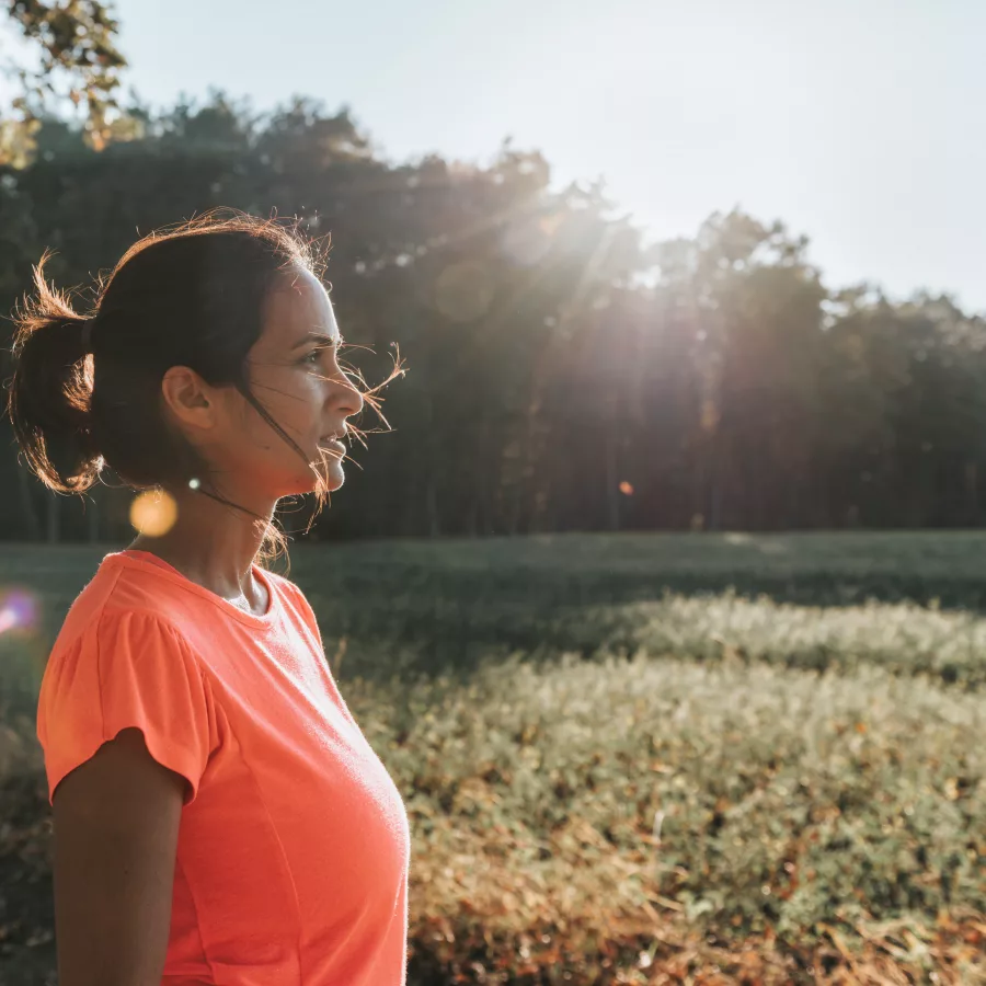 Eine Frau steht in einem offenen Feld und genießt die Natur. Sie trägt ein leuchtend orangefarbenes T-Shirt und hat ihre Haare zu einem lockeren Pferdeschwanz gebunden. Die Sonne scheint durch die Bäume im Hintergrund und erzeugt einen warmen Lichtschein, der die Szene idyllisch und friedlich wirken lässt. Die Umgebung ist grün und weitläufig, mit einem Wald im Hintergrund, der eine ruhige und entspannende Atmosphäre schafft.