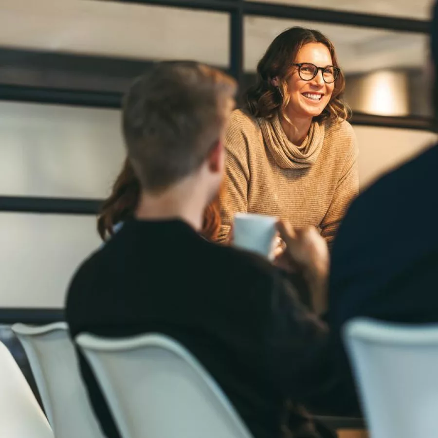 Das Bild zeigt drei Personen in einem modernen Büroraum, die miteinander sprechen. Zwei Männer sitzen an einem Konferenztisch, während die Frau am Tischende steht. Im Hintergrund sind große Fenster mit Milchglas zu sehen.