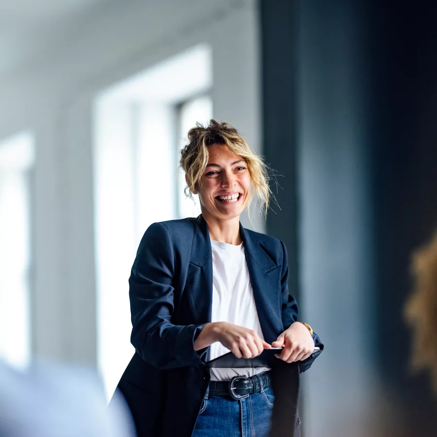 Eine Person hält einen Vortrag oder eine Präsentation vor mehreren Zuhörern in einem modern eingerichteten Raum. Sie trägt einen dunkelblauen Blazer, ein weißes Oberteil und eine Jeans. Die Atmosphäre wirkt professionell und aufmerksam.