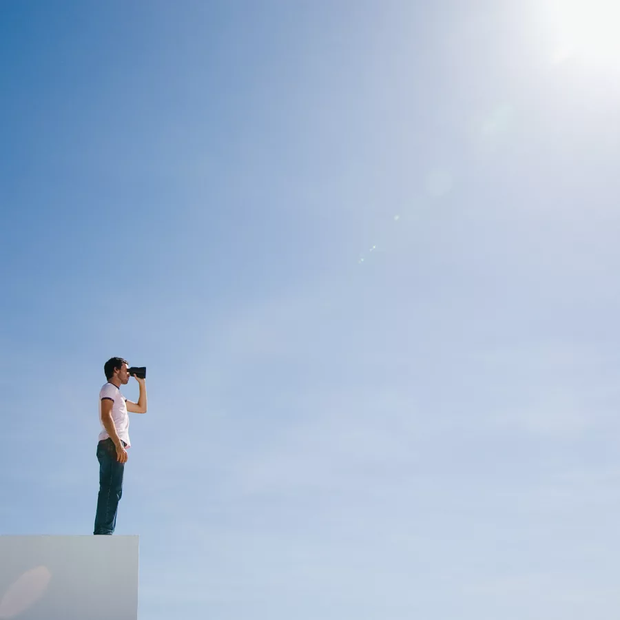 Eine Person steht auf einer weißen Kante und blickt mit einem Fernglas in den weiten, strahlend blauen Himmel. Die Szene vermittelt Weitblick, Neugier und den Wunsch, neue Horizonte zu erkunden. Die starke Sonne oben rechts verstärkt das Gefühl von Klarheit und Zukunftsorientierung.