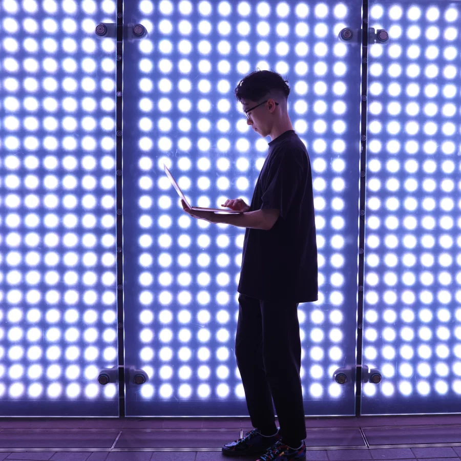 Eine Person steht seitlich vor einer leuchtenden Wand aus vielen LED-Lichtern und arbeitet an einem aufgeklappten Laptop. Das futuristisch wirkende Setting lässt den gesamten Hintergrund in hellem Weiß erstrahlen. Die dunkle Kleidung der Person hebt sich stark vom strahlenden Licht ab.