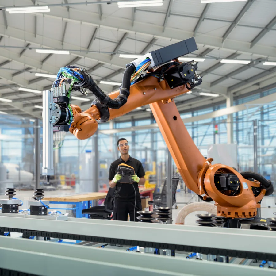 In einer modernen Fabrikhalle ist ein großer Industrieroboter im Einsatz. Im Hintergrund überwacht eine Person in Arbeitskleidung den Produktionsprozess über ein Steuergerät. Die Umgebung wirkt hell und technisch hochwertig, mit zahlreichen Glasabtrennungen und Maschinen.