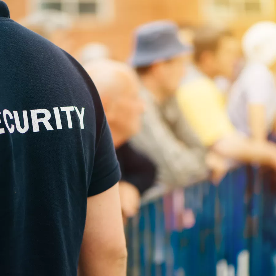 Das Bild zeigt einen Sicherheitsmitarbeiter, der ein schwarzes T-Shirt mit der Aufschrift "SECURITY" trägt. Der Sicherheitsmitarbeiter steht im Vordergrund und überwacht eine Menschenmenge, die hinter einer blauen Absperrung steht. Die Szene scheint bei Tageslicht stattzufinden, und im Hintergrund sind mehrere Personen zu sehen, die sich an der Absperrung aufhalten. Das Bild vermittelt ein Gefühl von Sicherheit und Ordnung bei einer Veranstaltung oder Versammlung.
