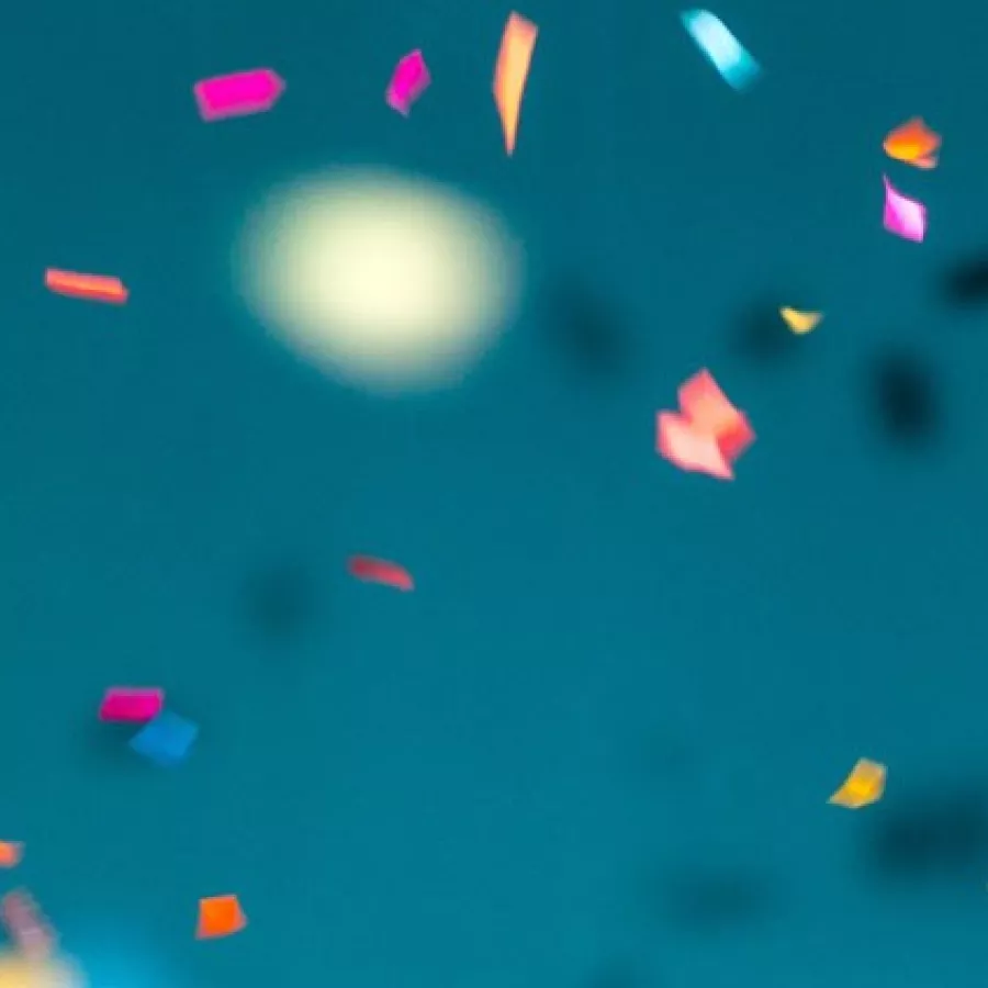 Das Bild zeigt eine Szene mit buntem Konfetti, das vor einem blauen Hintergrund in der Luft schwebt. Die Konfettistücke sind in verschiedenen Farben wie Pink, Gelb, Blau, Grün und Orange gehalten und scheinen in Bewegung zu sein, als ob sie gerade geworfen wurden. Der Hintergrund ist einfarbig blau und vermittelt einen festlichen und fröhlichen Eindruck. Die Szene könnte Teil einer Feier oder eines besonderen Anlasses sein.