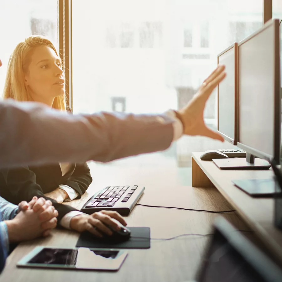 Drei Personen arbeiten an Computern in einem Büro, eine Person zeigt auf einen der Bildschirme.