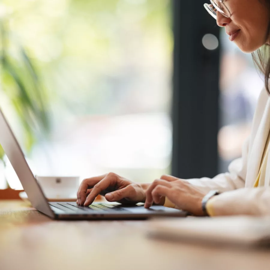 Das Bild zeigt eine Person, die an einem Laptop arbeitet. Die Person trägt eine Brille und ein weißes Oberteil. Sie sitzt an einem Holztisch und tippt auf der Tastatur des Laptops. Im Hintergrund sind unscharf grüne Pflanzen und ein Fenster zu erkennen, durch das natürliches Licht hereinfällt. Die Szene wirkt ruhig und konzentriert.