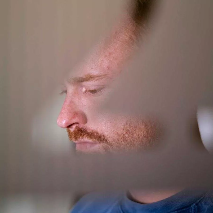 Das Bild zeigt eine Person, die ein blaues Oberteil trägt. Der Hintergrund ist unscharf, aber es scheint sich um einen Innenraum zu handeln, möglicherweise ein Zimmer mit hellen Wänden. Ein Teil eines Möbelstücks oder einer Wanddekoration ist ebenfalls im Hintergrund zu erkennen. Die Beleuchtung ist weich und natürlich.